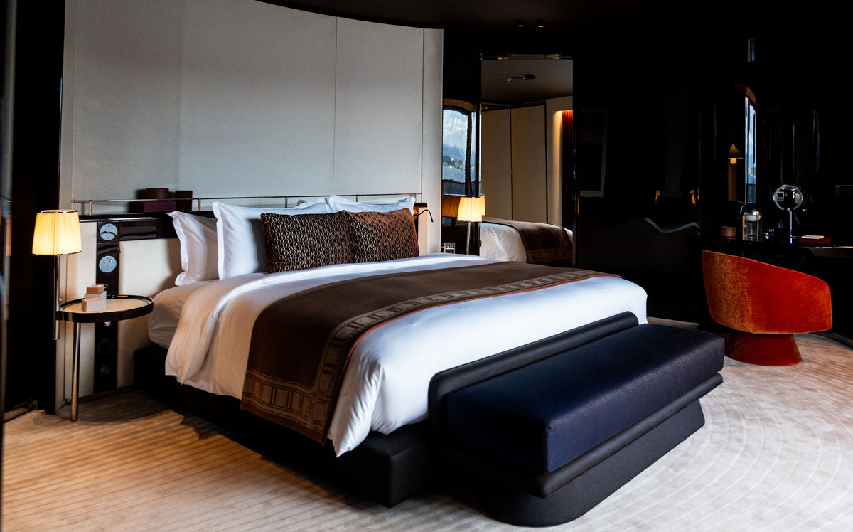 A luxury modern bedroom featuring a large bed dressed in crisp white linens with dark brown patterned accents, set against a dark wall with bedside tables and lamps. A dark upholstered bench sits at the foot of the bed, and a vibrant orange-red accent chair is visible in the background.